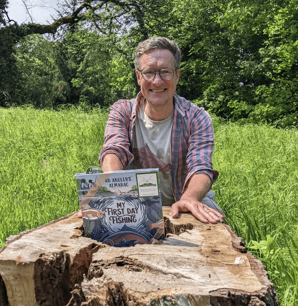 Will Millard kneeling at a tree stump holding a copy of My First Day Fishing