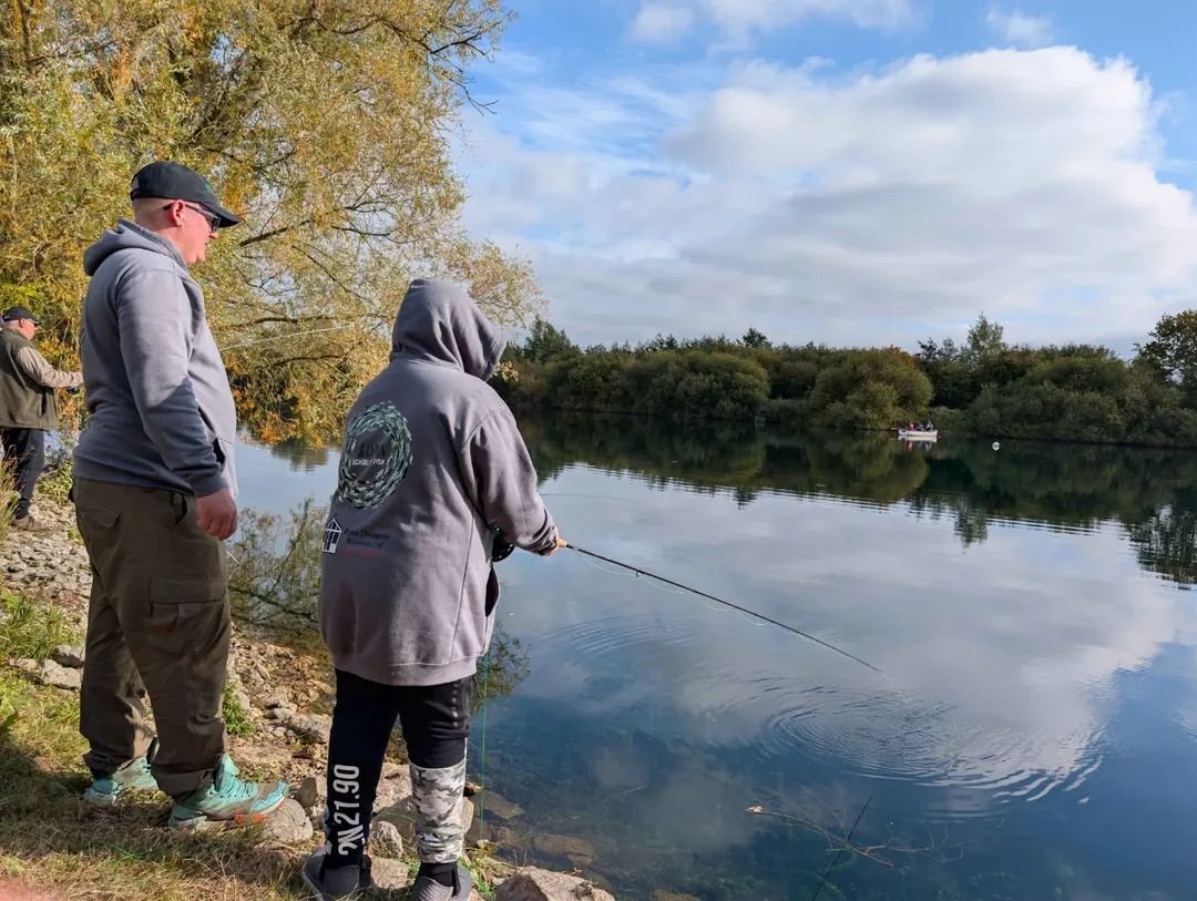 A School Of Fish coach stands beside a young angler fishing a flat calm lake from the bank